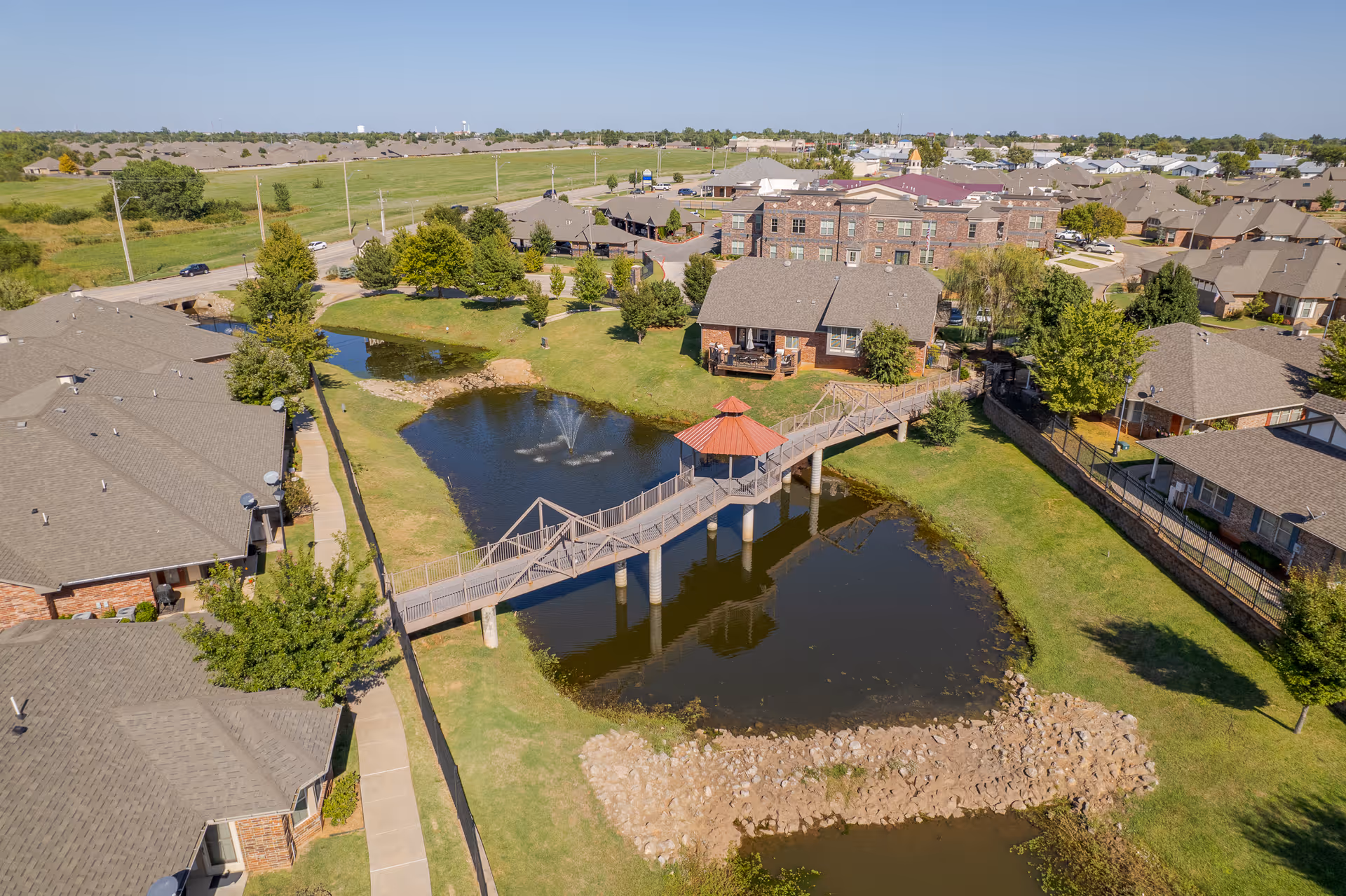 Aerial view of a senior living facility named Grace Pointe featuring multiple residential buildings surrounding a pond with a fountain. A wooden bridge with a small covered gazebo crosses the pond, connecting the buildings. The area is landscaped with green grass, trees, and paved walkways under a clear blue sky.