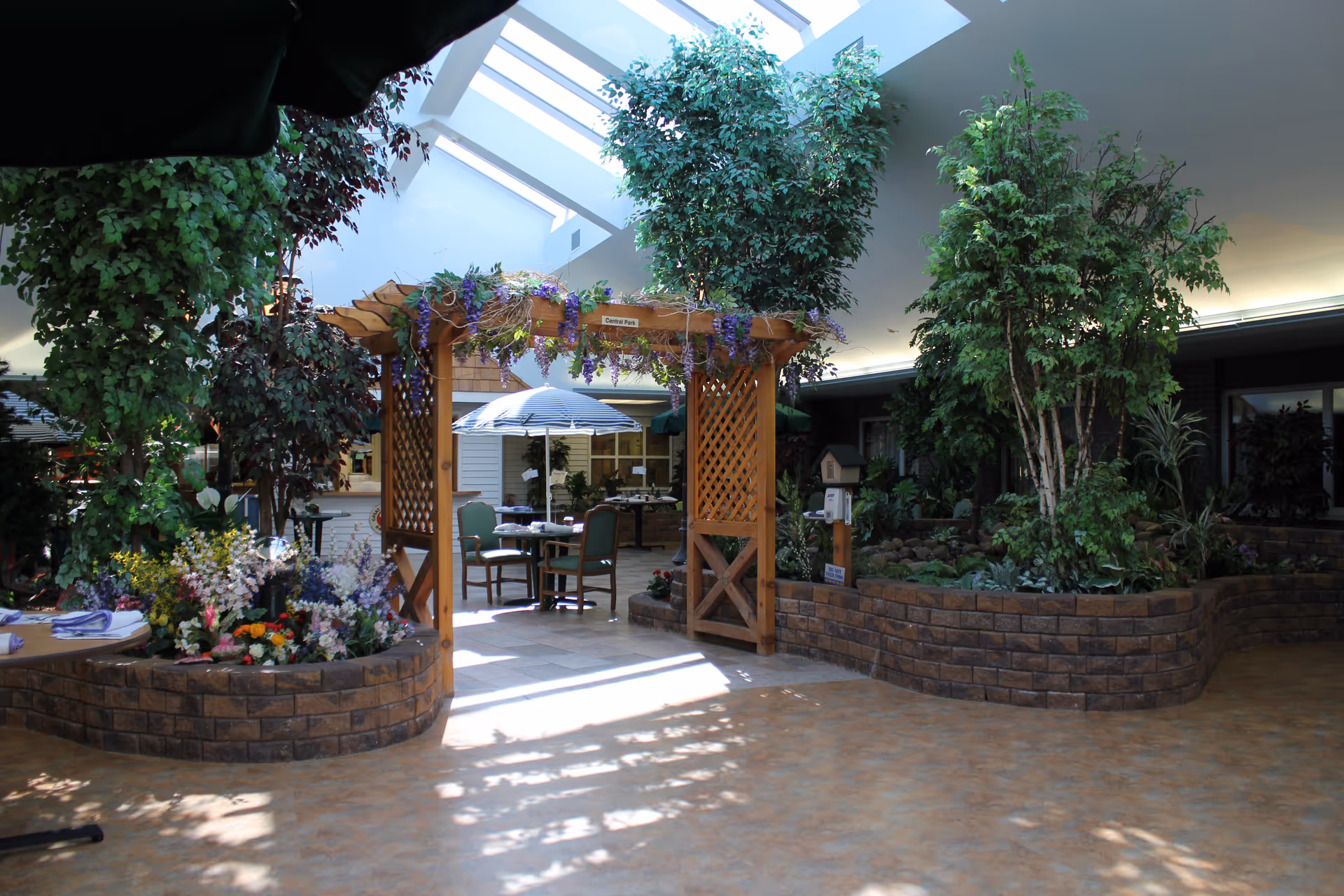 Indoor atrium with a wooden pergola, abundant plants, skylights, and seating with tables and umbrellas.