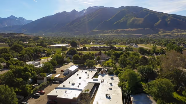 Aerial view of a facility with a flat white roof surrounded by trees and residential buildings, with a mountain range in the background under a clear sky.