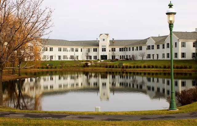 A large white two-story building with multiple windows is reflected in a calm pond in the foreground. There is a green lamppost on the right side near the water, and some leafless trees and grass surround the pond.