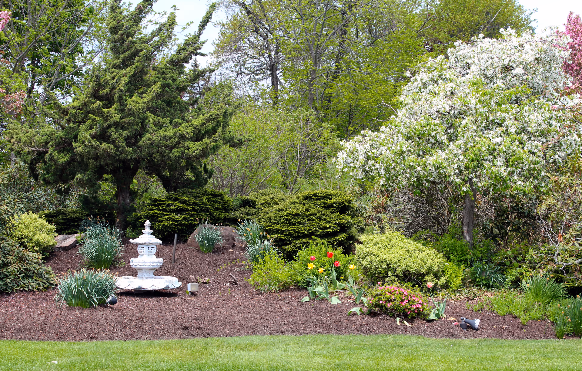 Landscaped garden bed with a white decorative fountain, flowering shrubs, and green trees.
