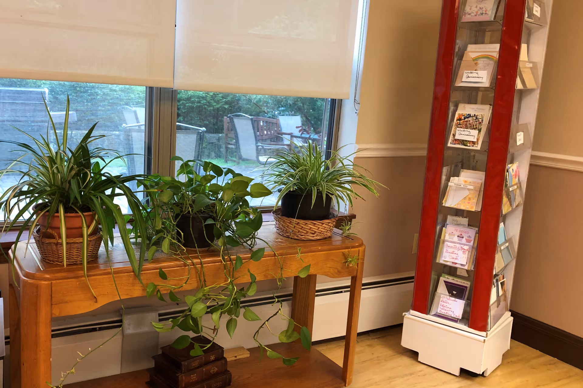 Indoor scene with a wooden table holding three potted green plants placed in front of a window with white roller blinds. Outside the window, patio chairs and a bench are visible. To the right of the table is a tall, red and white rotating card display rack filled with various greeting cards. The room has beige walls with white trim and a wooden floor.