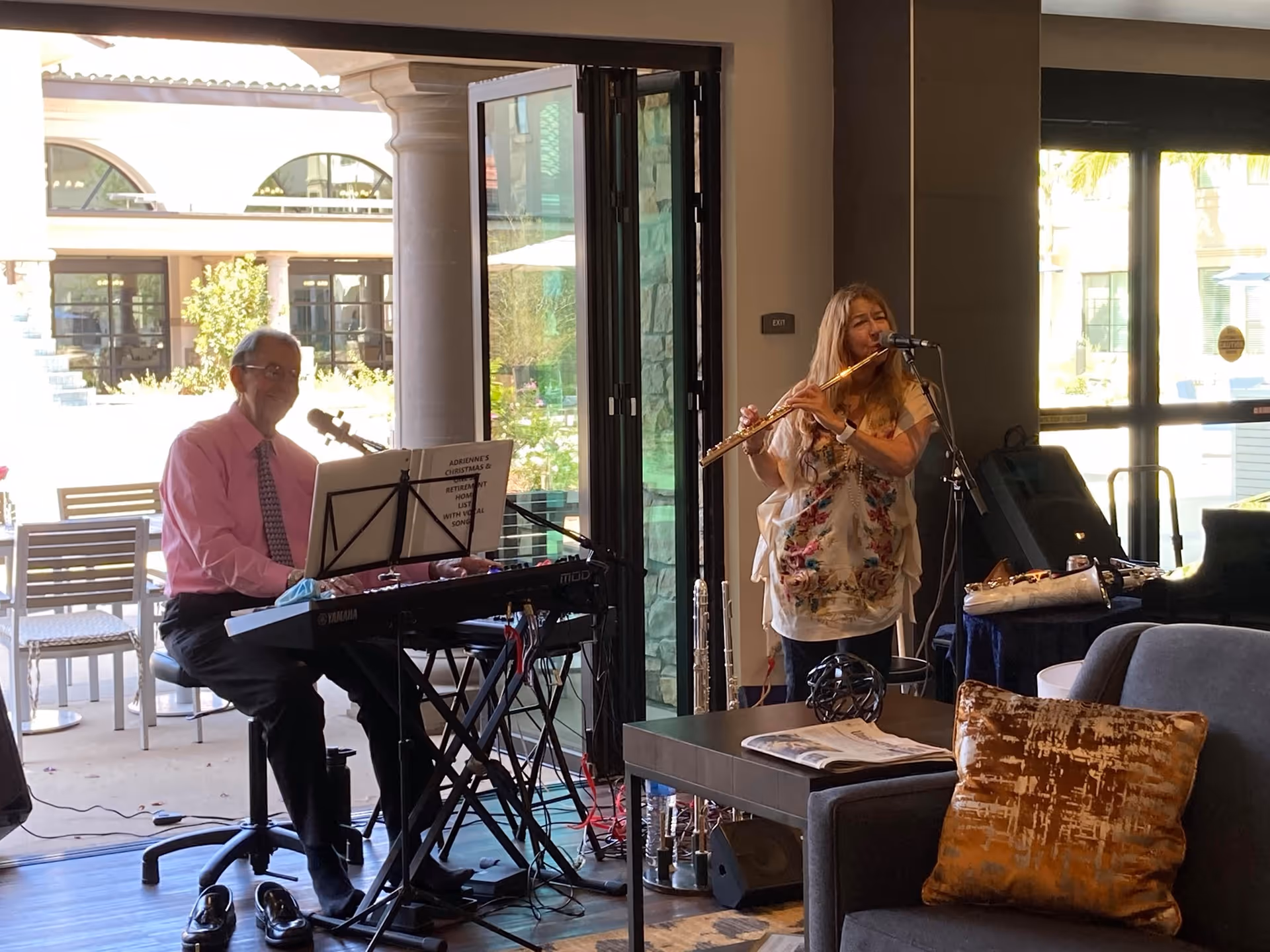 Two musicians, a keyboard player and a flutist, performing in a lounge/common area with seating and open patio doors.