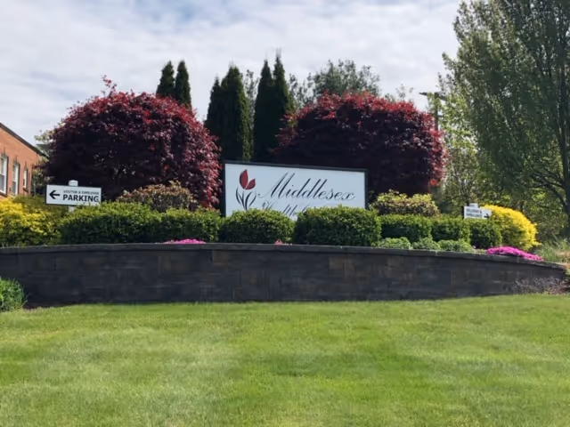 A landscaped garden area with a stone retaining wall and various bushes and trees. In the center is a sign that reads 'Middlesex Health Care Center' with a red tulip logo. There are directional signs on either side of the garden indicating parking and entrance directions. The sky is partly cloudy.