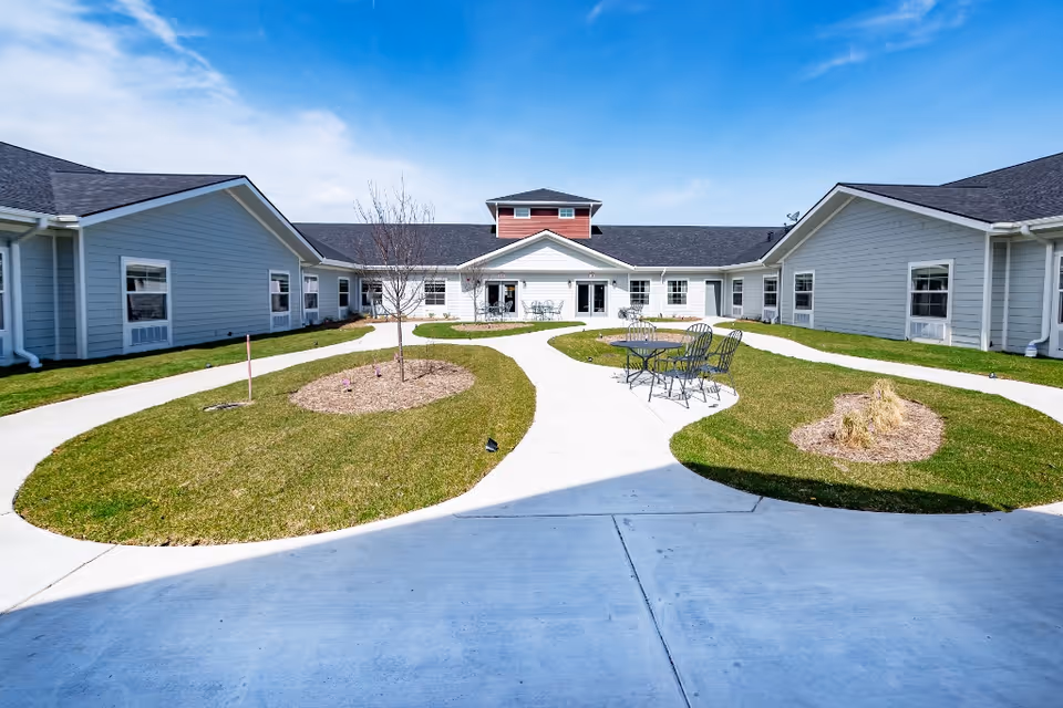 Outdoor courtyard area of a senior living facility with a concrete pathway winding through green grass and small landscaped areas. There are several metal tables and chairs placed on the grass near the pathway. The building surrounding the courtyard is single-story with gray siding and white trim under a blue sky with some clouds.