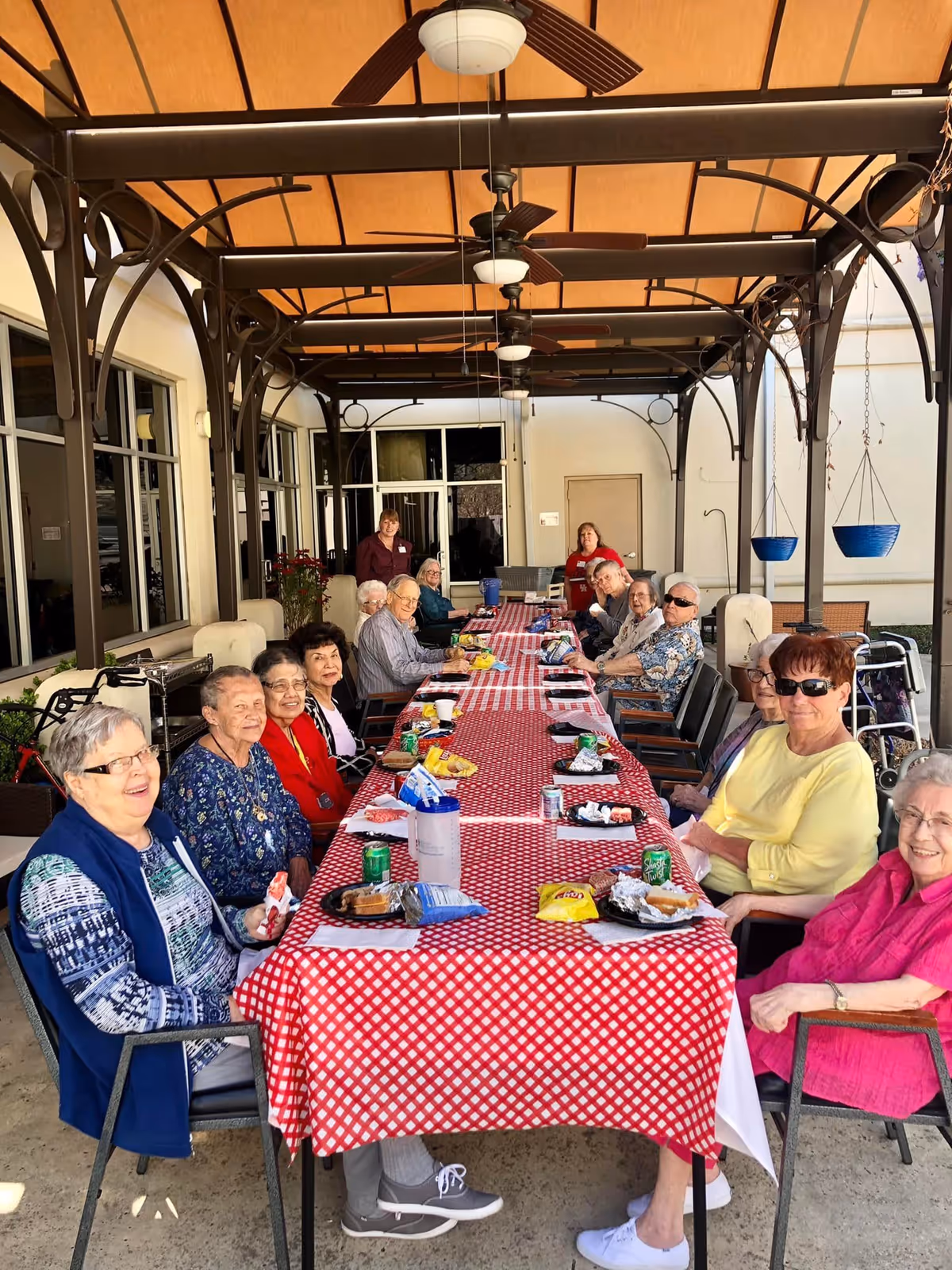A group of elderly people sitting around a long table covered with a red and white checkered tablecloth under a covered outdoor patio. They are enjoying snacks and drinks, with some smiling and looking at the camera. The patio has ceiling fans and hanging planters, and two staff members are standing in the background near the building entrance.