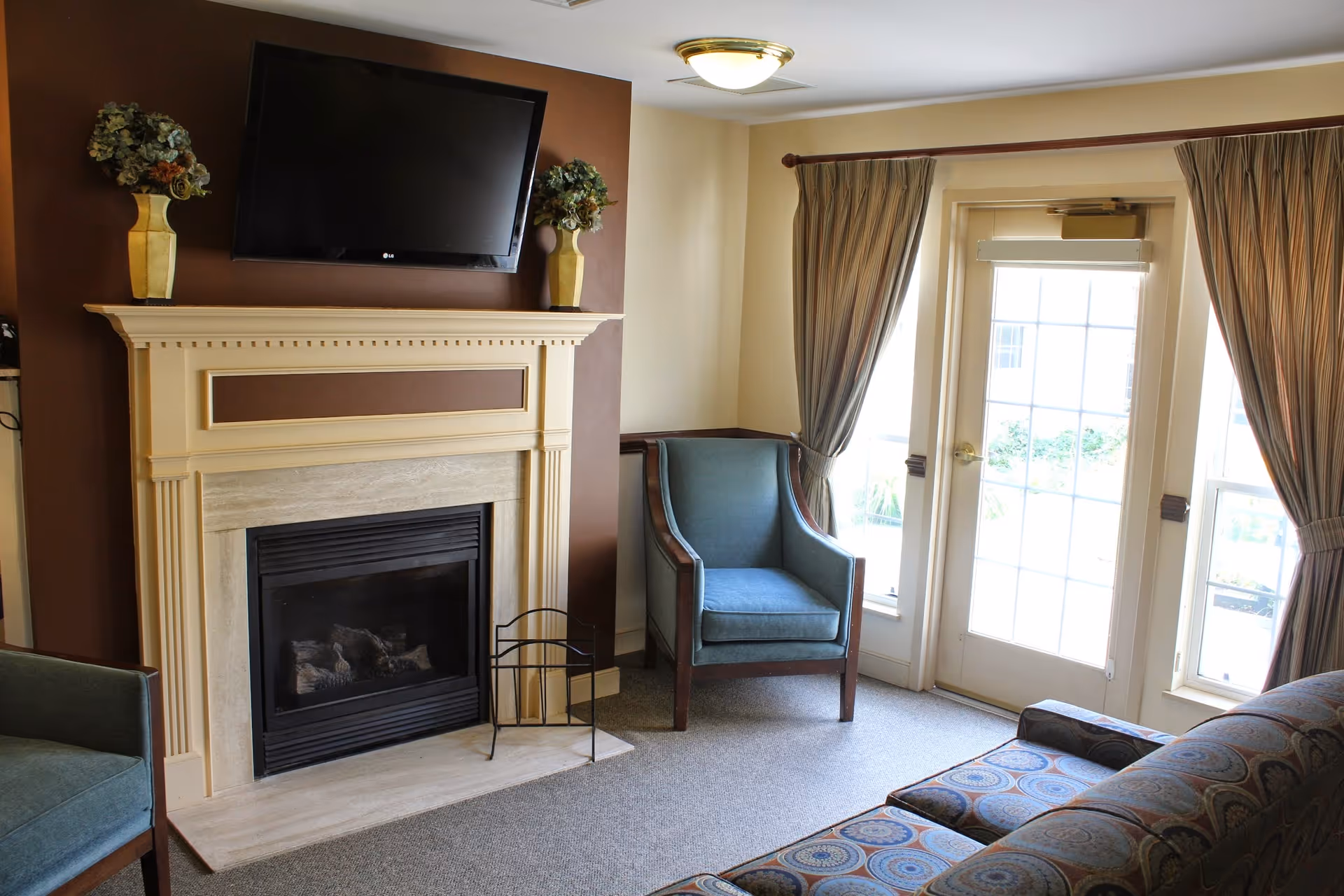 A cozy living room area with a fireplace and a mounted flat-screen TV above it. Two vases with floral arrangements are placed on the mantel. There are two blue armchairs and a patterned sofa arranged around the fireplace. Large windows with curtains and a glass door allow natural light into the room.
