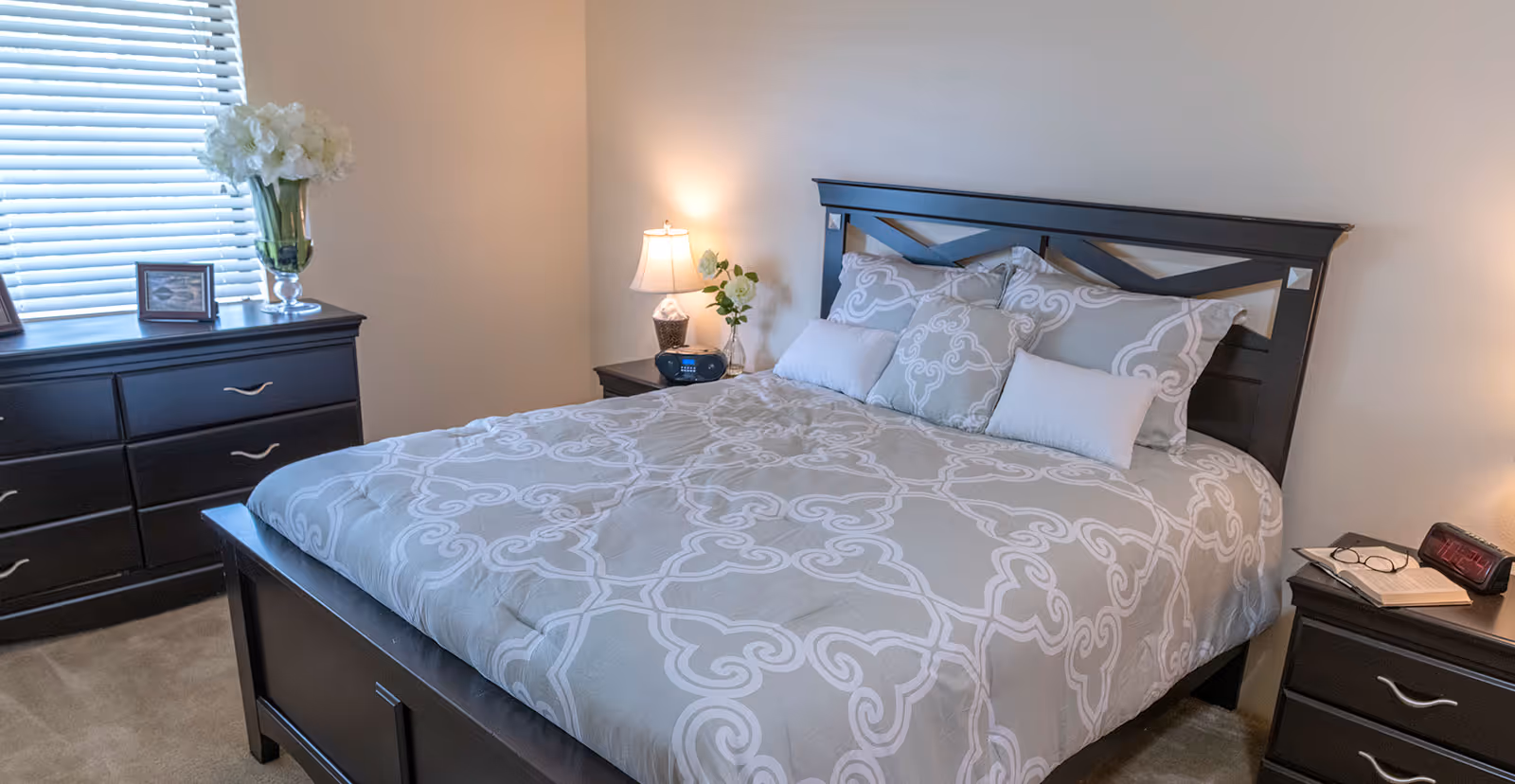 A neatly made bed with a patterned gray and white comforter and multiple pillows in a bedroom. The room features dark wood furniture including a dresser with a vase of white flowers and framed pictures, and two nightstands with a lamp, alarm clock, and a book with glasses. A window with closed blinds is visible behind the dresser.