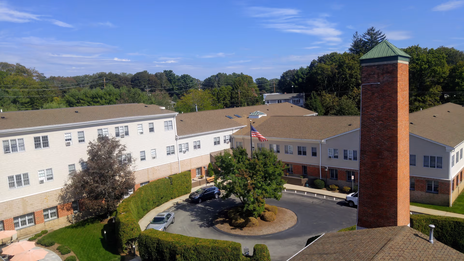 Aerial view of a large assisted living and skilled nursing community building with beige siding and brick accents, surrounded by trees and greenery. There is a circular driveway with a flagpole in the center and several parked cars. The building has multiple windows and a tall brick chimney with a green metal cap.