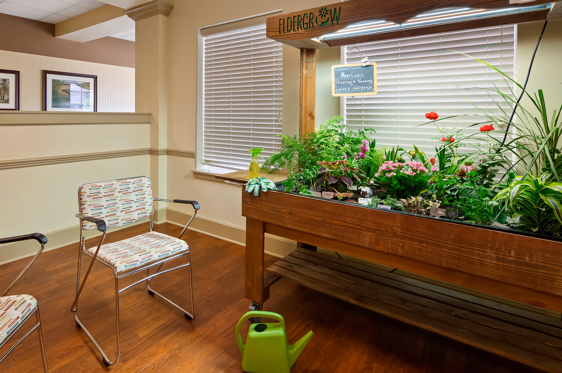 Indoor gardening station with a raised wooden planter filled with plants and flowers, two chairs, and a green watering can in a senior living common area.