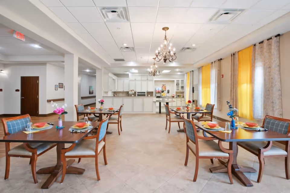 A bright dining room with several wooden tables and chairs arranged neatly. Each table is set with plates, bowls, glasses, and napkins, and decorated with small vases of colorful flowers. The room has large windows with yellow and patterned curtains, and two ornate chandeliers hanging from the ceiling. In the background, there is a kitchen area with white cabinets and appliances.