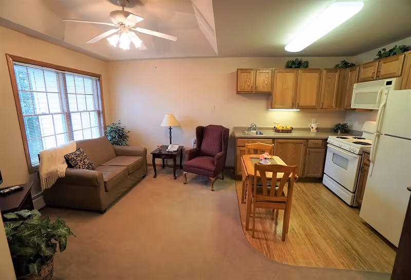 A cozy living area with a beige sofa, a purple armchair, a wooden side table with a lamp, and a small dining table with two chairs. The room has a large window with blinds, a ceiling fan with lights, and an open kitchen with wooden cabinets, a white refrigerator, stove, microwave, and a sink. The floor transitions from carpet in the living area to wood in the kitchen.
