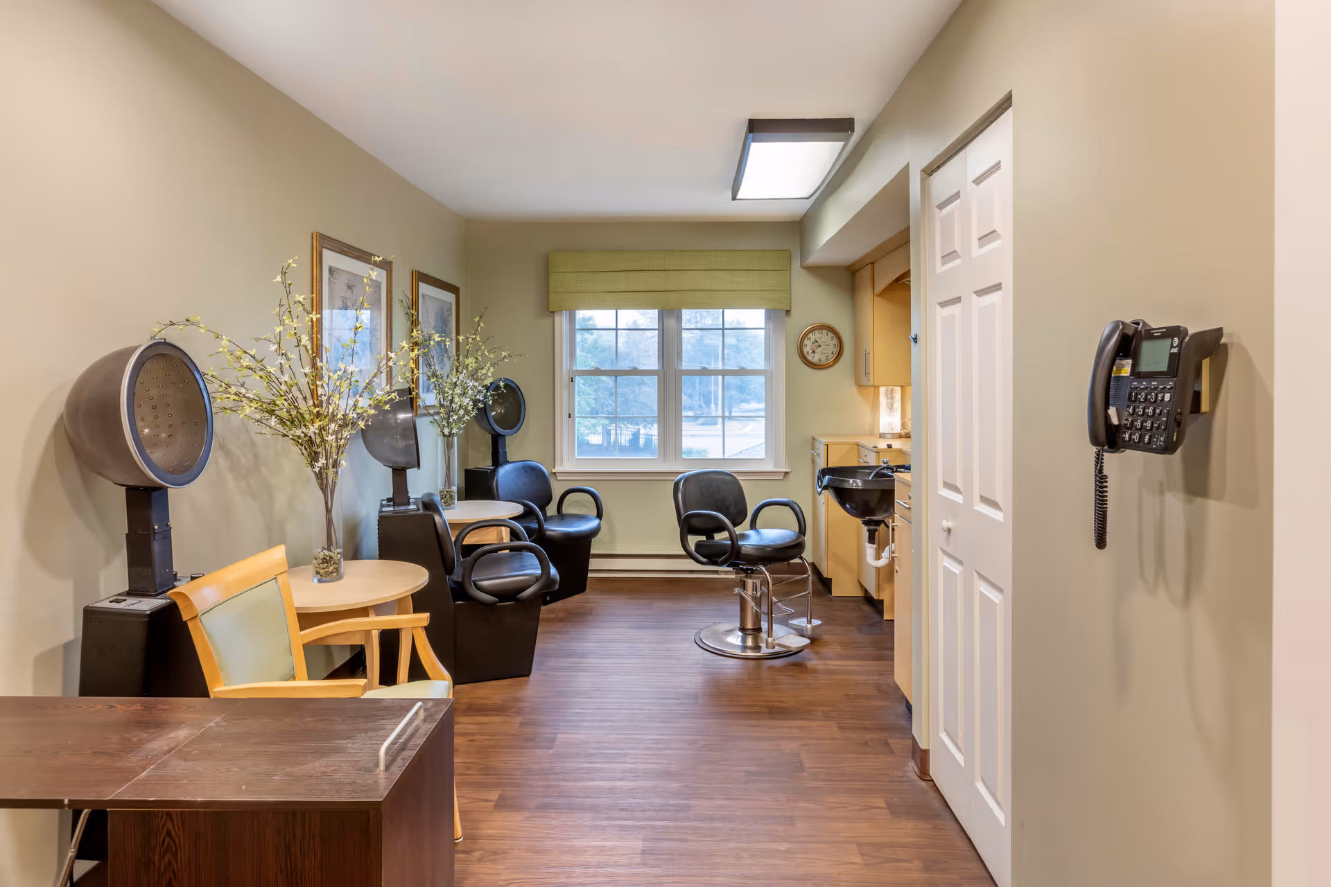 Interior view of a salon area in an assisted living facility with salon chairs, hair dryers, a small table with a vase of flowers, framed pictures on the wall, a window with a green valance, and a wall-mounted telephone.