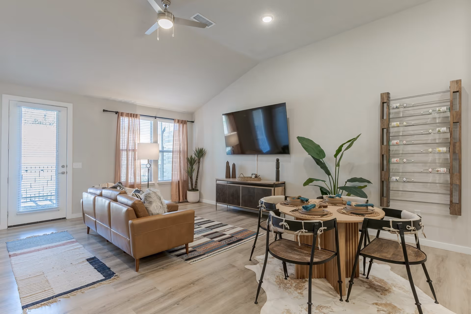 A modern living and dining area with a brown leather sofa, a round wooden dining table set with four chairs, a large flat-screen TV mounted on the wall, a floor lamp, potted plants, and large windows with sheer curtains letting in natural light.