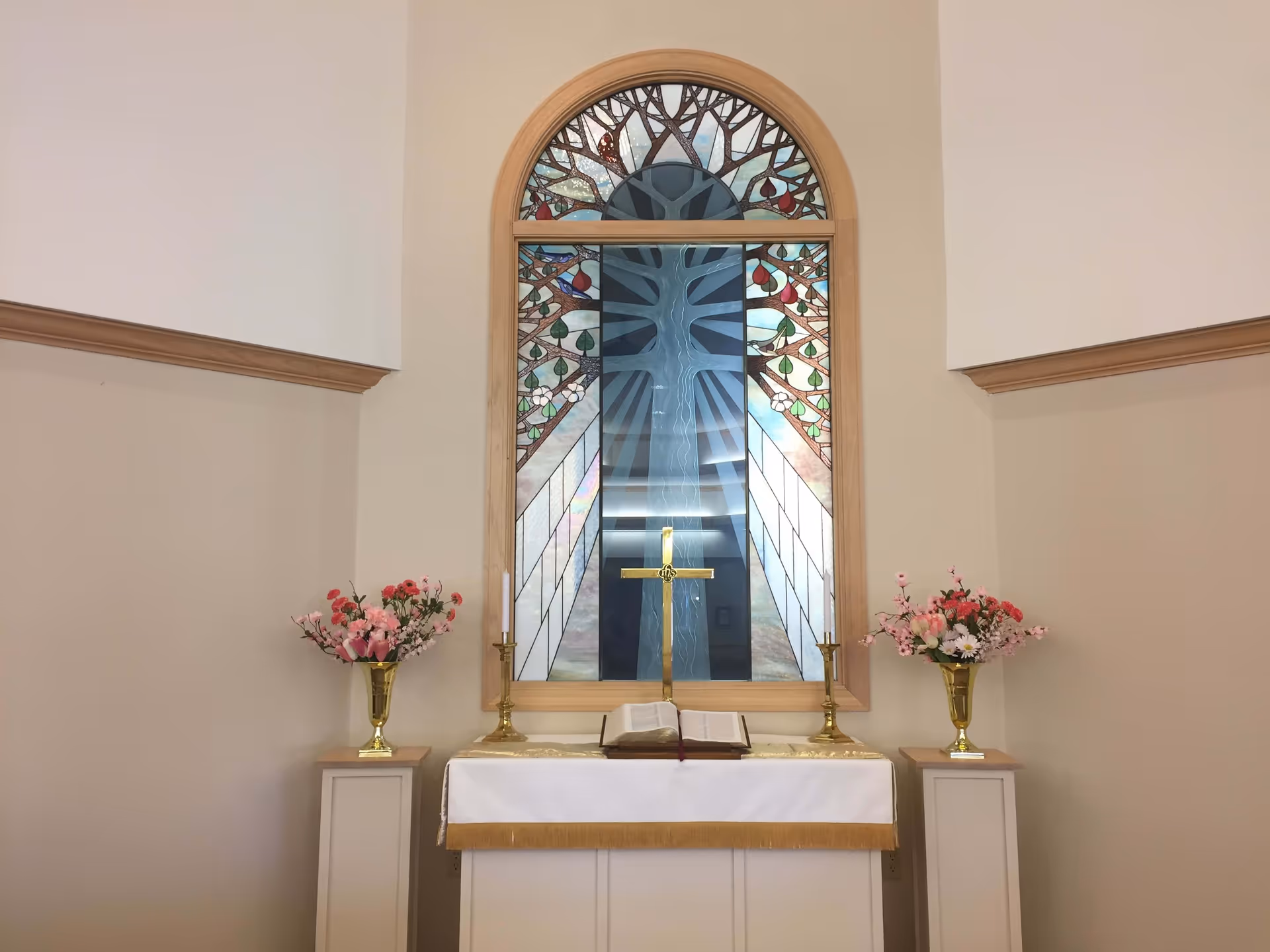 Interior view of a small chapel or prayer room with a stained glass window depicting a tree behind an altar. The altar is covered with a white cloth with gold trim and holds an open Bible, a gold cross, two gold candlesticks with white candles, and two gold vases with pink and red flowers on either side.