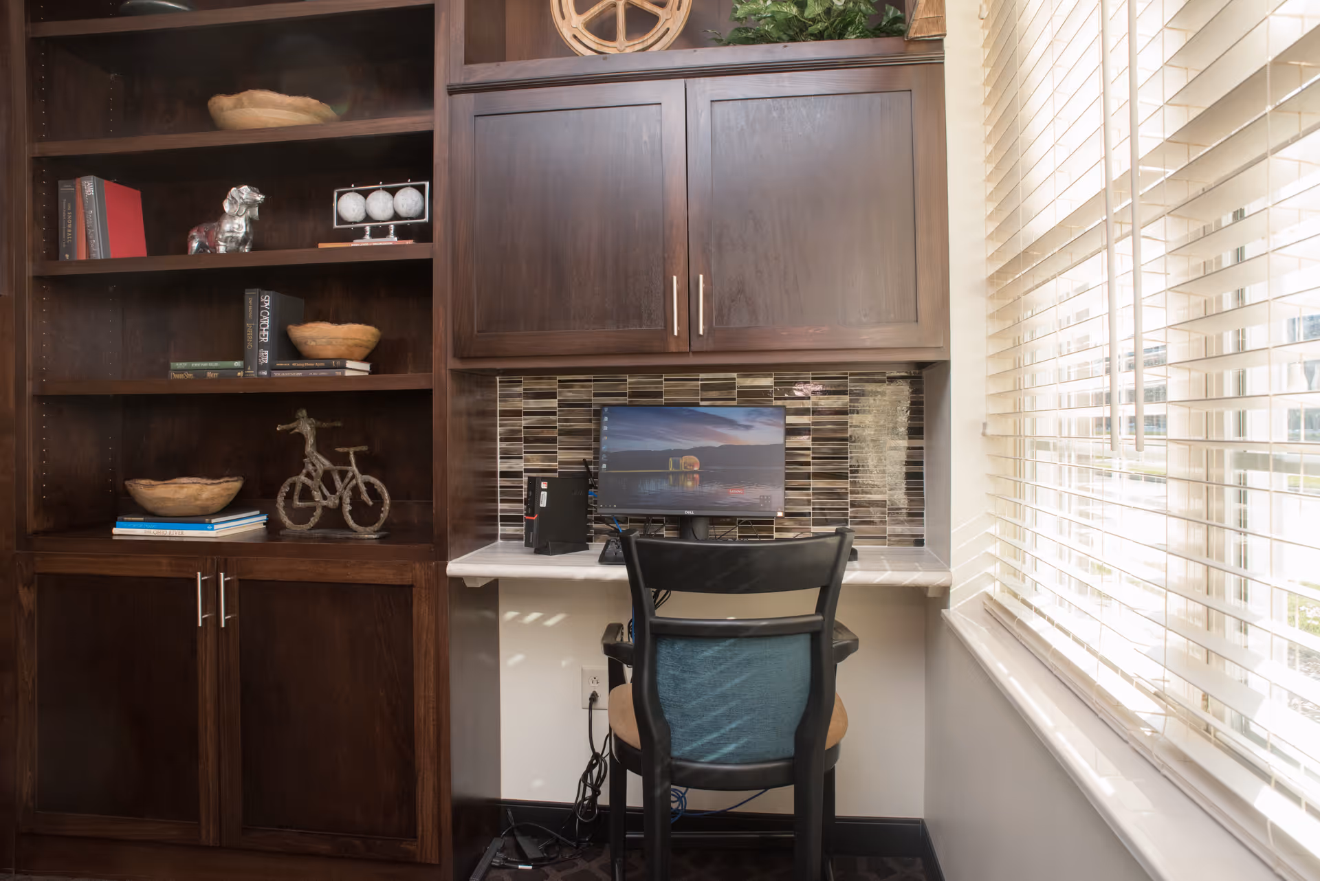 A small workspace with a computer monitor on a built-in desk, a black chair, and dark wooden cabinets and shelves. The shelves hold decorative items including books, a small bicycle sculpture, and bowls. A window with white blinds is on the right, letting in natural light.