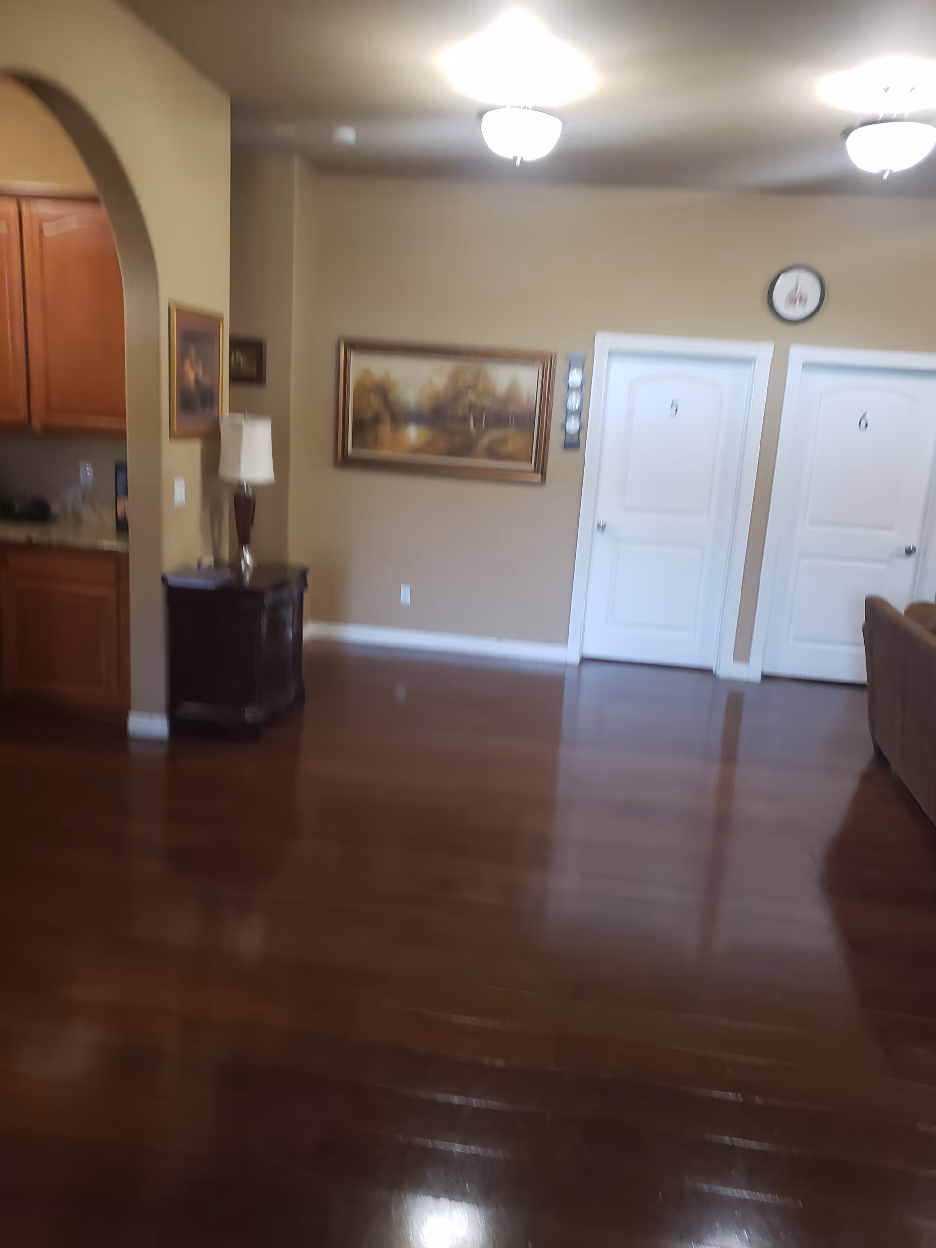 Interior view of a senior living facility showing a polished wooden floor, two white doors labeled 5 and 6, a brown couch partially visible on the right, a small dark wooden table with a lamp and some items on it, a kitchen area with wooden cabinets on the left, beige walls with framed artwork, and ceiling lights.