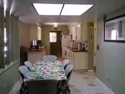 Narrow dining-kitchen room with a long table covered by a floral tablecloth surrounded by chairs, with cabinets, a refrigerator and microwave along the far wall.