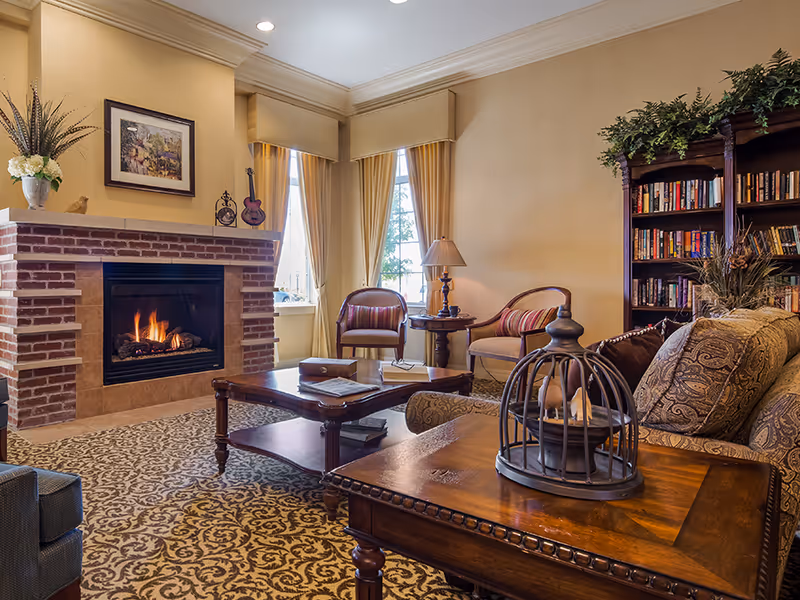 Cozy living room with a lit brick fireplace, armchairs, coffee table, and a bookshelf in warm decor.