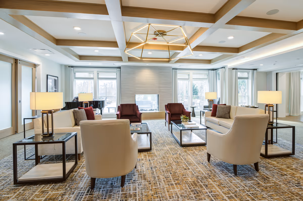 Light-filled communal lounge with upholstered sofas and armchairs arranged around coffee tables and a central fireplace.