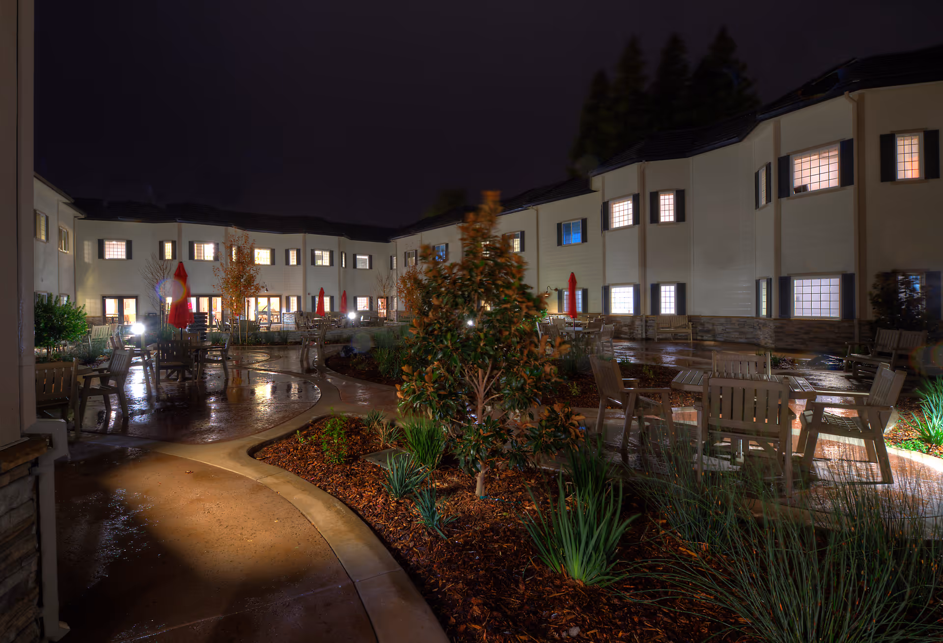 Night view of a wet courtyard with patio tables and chairs surrounded by a two-story senior living building with lit windows.