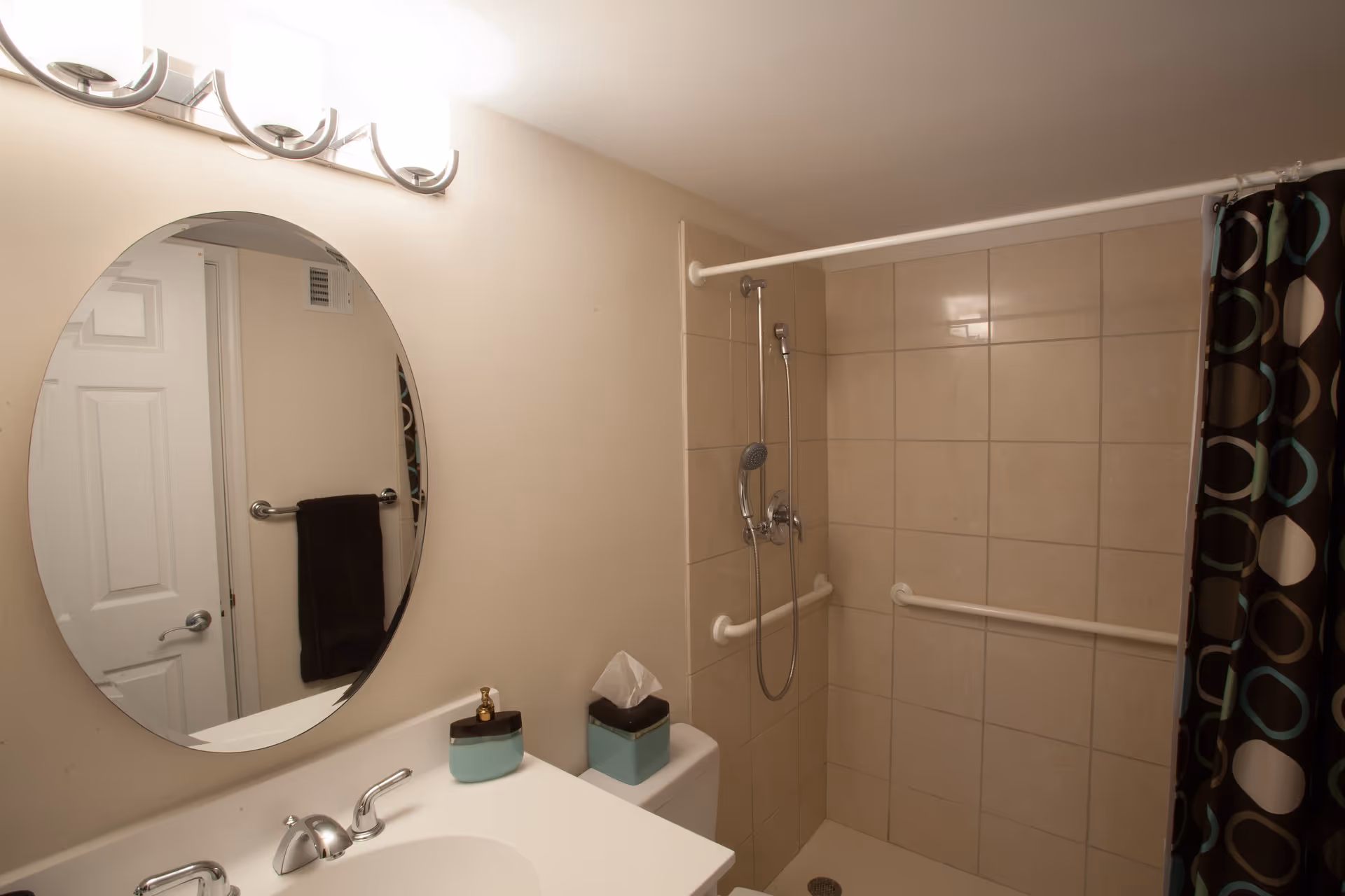 A bathroom with a white sink and countertop, a round mirror above the sink, a towel hanging on a rack next to a white door, a tiled shower area with grab bars and a handheld showerhead, and a shower curtain with a circular pattern.