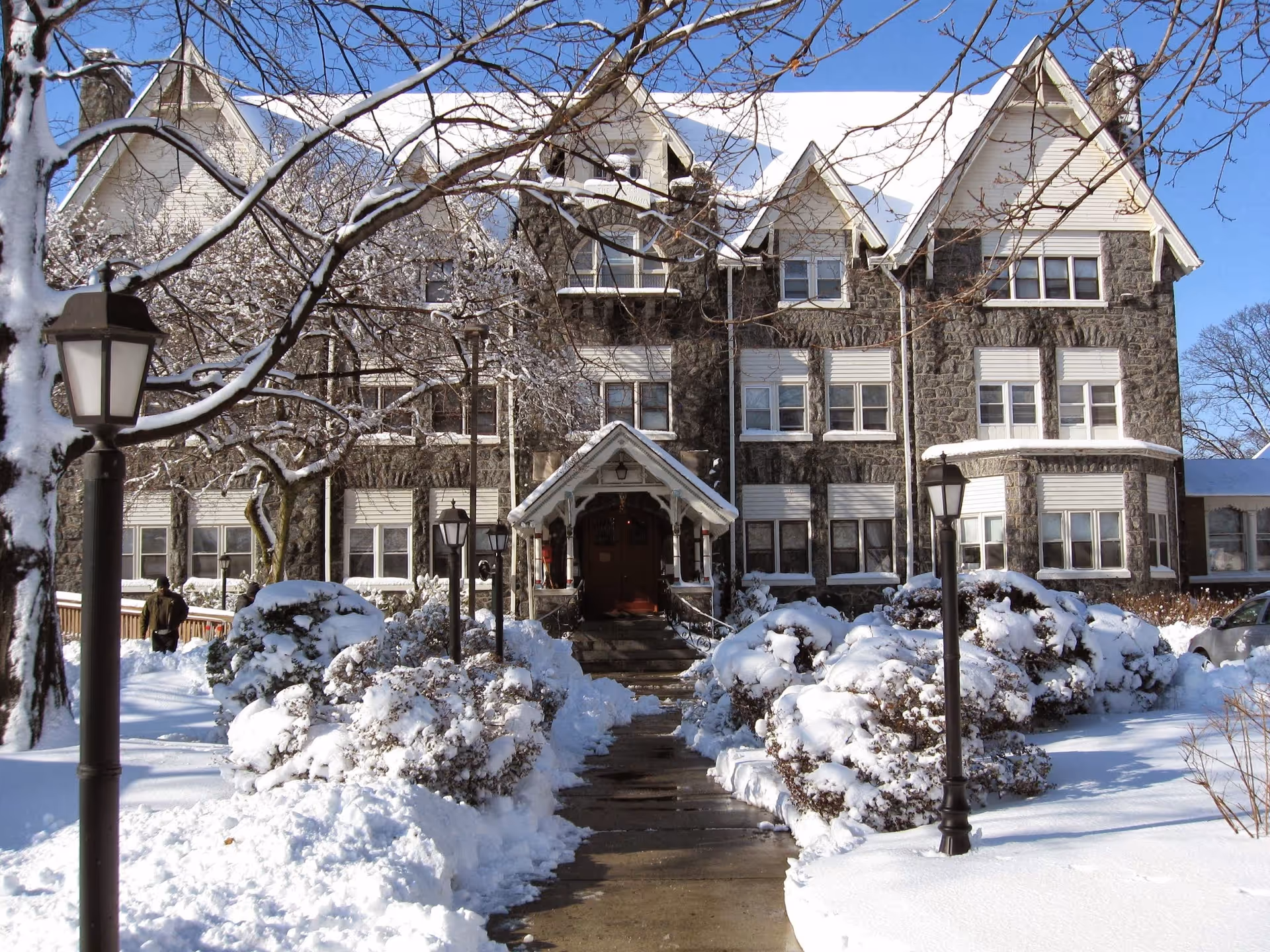 A large stone building with white trim and multiple windows, covered in snow. A pathway lined with snow-covered bushes and lamp posts leads to a covered entrance. Bare tree branches are visible, and the sky is clear and blue.