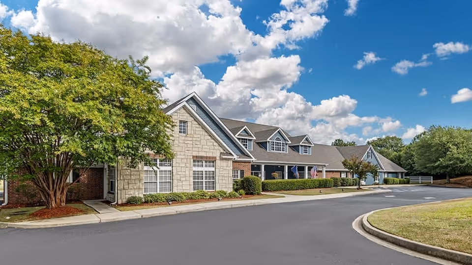 Exterior view of a senior living facility building with a combination of brick and stone facade, surrounded by trees and shrubs under a partly cloudy blue sky.