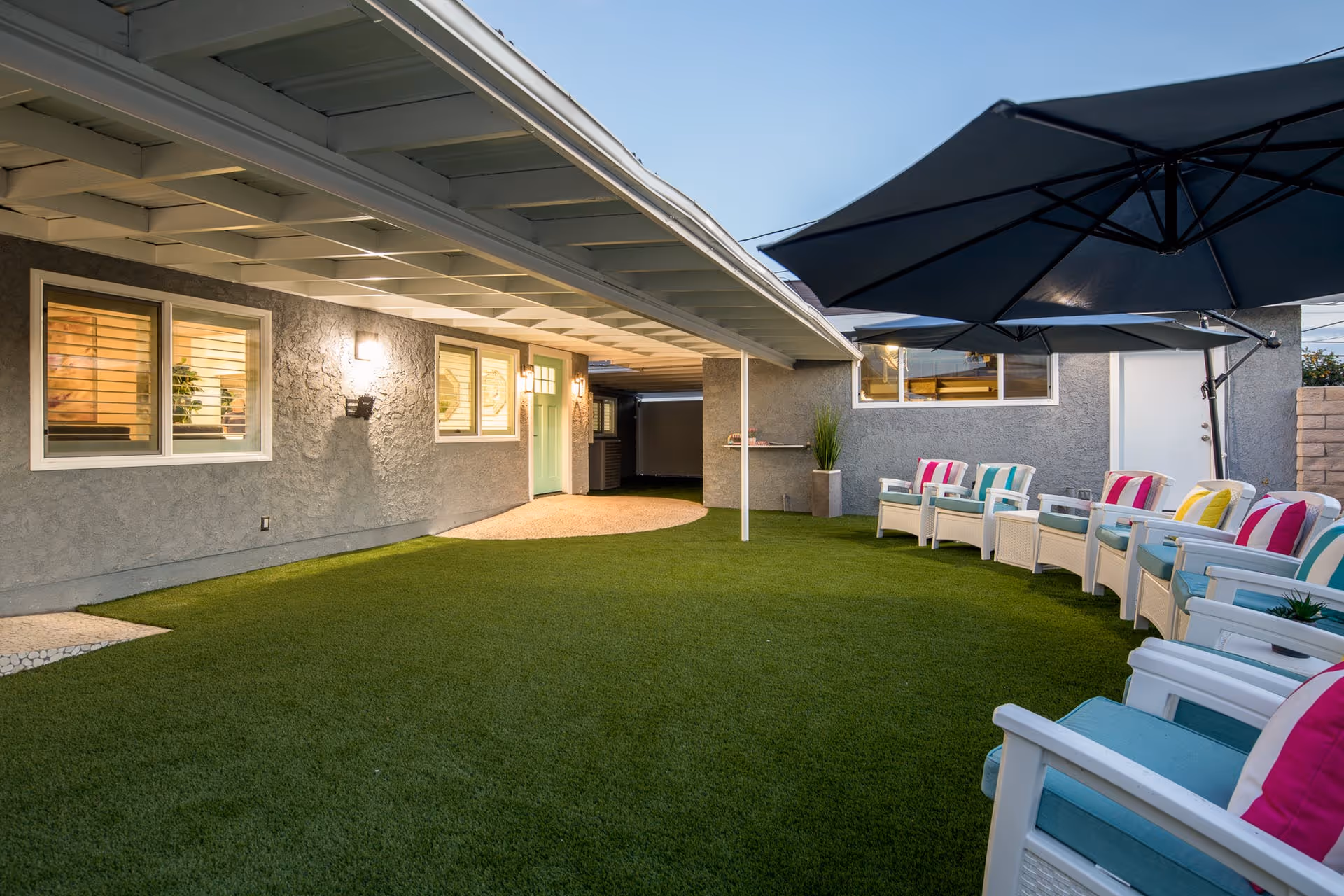 Outdoor patio area with artificial grass, white chairs with colorful cushions arranged in a row under large black umbrellas, and a gray building with windows and a green door in the background.