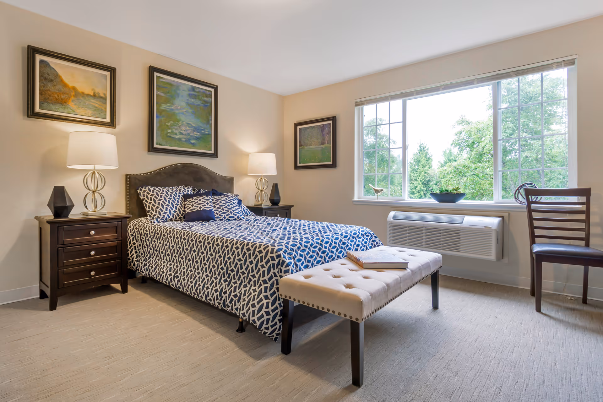 A bright bedroom with a bed covered in a blue and white patterned comforter, two matching pillows, and a small decorative pillow. There are two dark wooden nightstands on either side of the bed, each with a modern lamp and decorative items. Above the bed are three framed paintings. A large window with white blinds lets in natural light and shows green trees outside. In front of the bed is a beige tufted bench with a book on it, and to the right is a wooden chair.