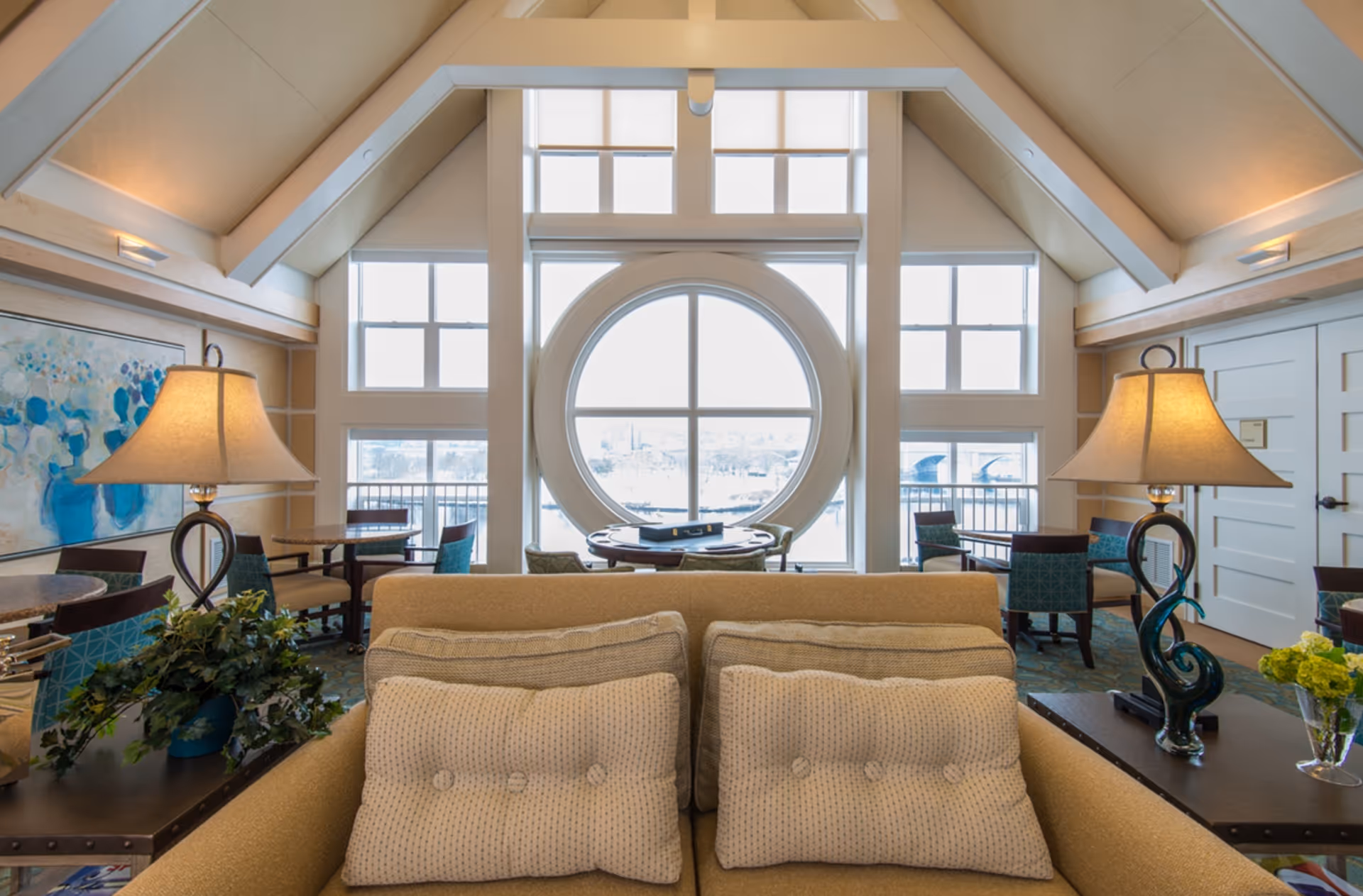 A bright and cozy living room area with a beige sofa adorned with two patterned cushions. Behind the sofa are two side tables, each with a decorative lamp and plants. The room features a large circular window flanked by rectangular windows, allowing natural light to fill the space. Several chairs and small tables are arranged near the windows, and the ceiling has exposed beams with soft lighting.