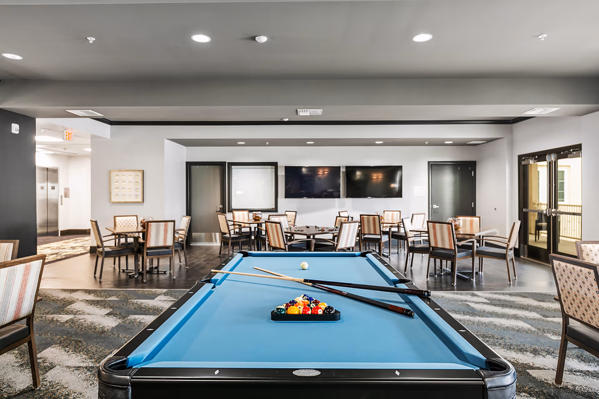 Indoor community game room with a blue pool table in the foreground and tables and chairs arranged in the background.
