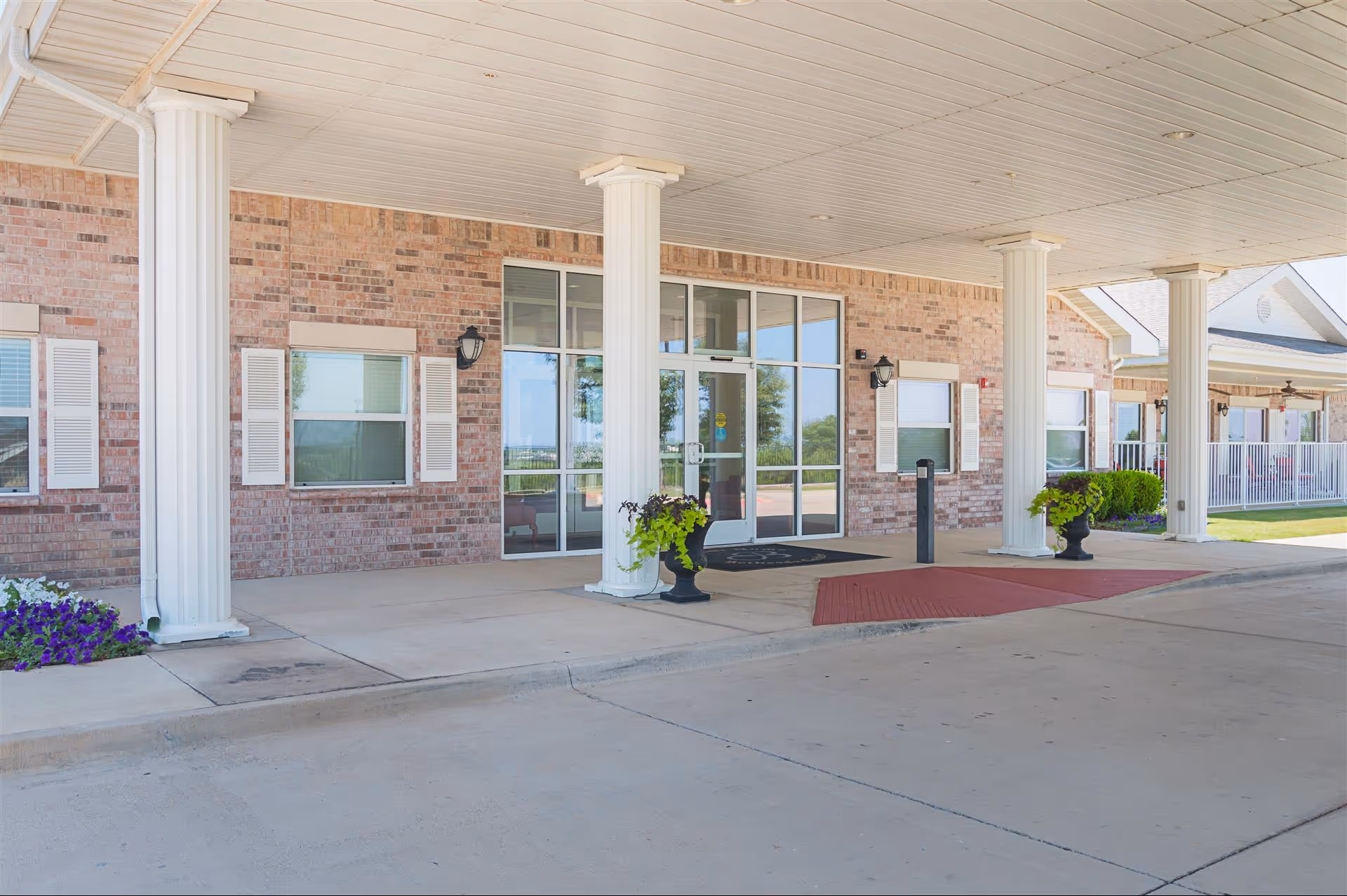 Entrance of a senior living facility with large white columns supporting a covered driveway. The building has a brick exterior with multiple windows and glass doors. There are potted plants near the columns and a red textured mat on the ground near the entrance.