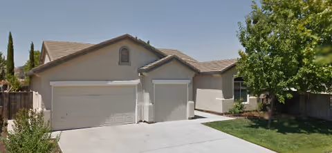 Single-story residential building with a two-car garage and a driveway. The house has a beige exterior with a tiled roof. There is a green lawn and a tree on the right side of the driveway, and a wooden fence on both sides of the property.