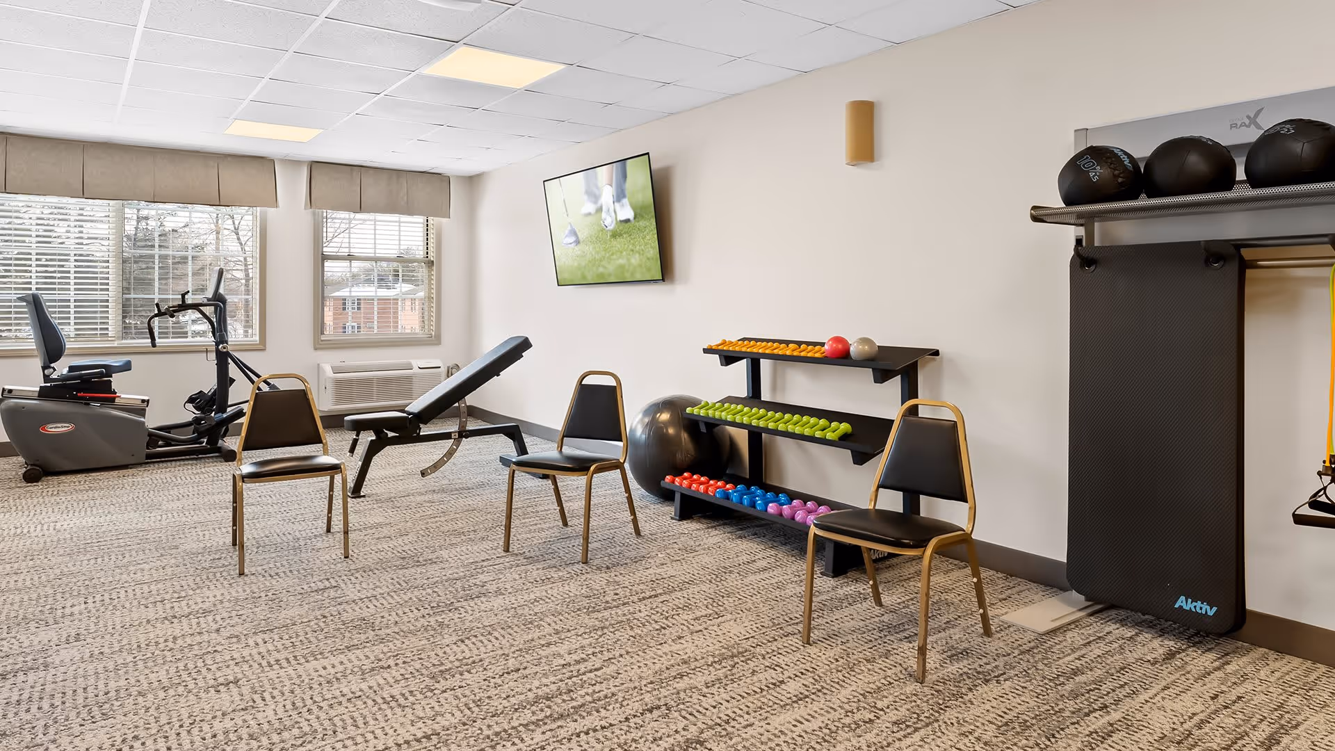 A well-lit exercise room with two windows, a recumbent exercise bike, an adjustable weight bench, three chairs, a rack of colorful dumbbells, a large exercise ball, a black exercise mat hanging on the wall, and a wall-mounted TV displaying a golf scene.