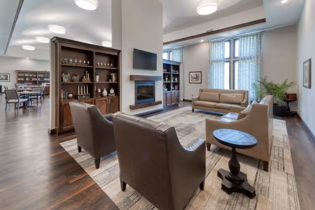 A spacious and well-lit living room area with large windows covered by sheer curtains. The room features a modern fireplace mounted on a central column, a wall-mounted flat-screen TV above it, and built-in wooden shelves with decorative items. There are two brown leather armchairs, a beige sofa, and a beige loveseat arranged around a patterned area rug. The room has dark wood flooring and neutral-colored walls, creating a warm and inviting atmosphere.