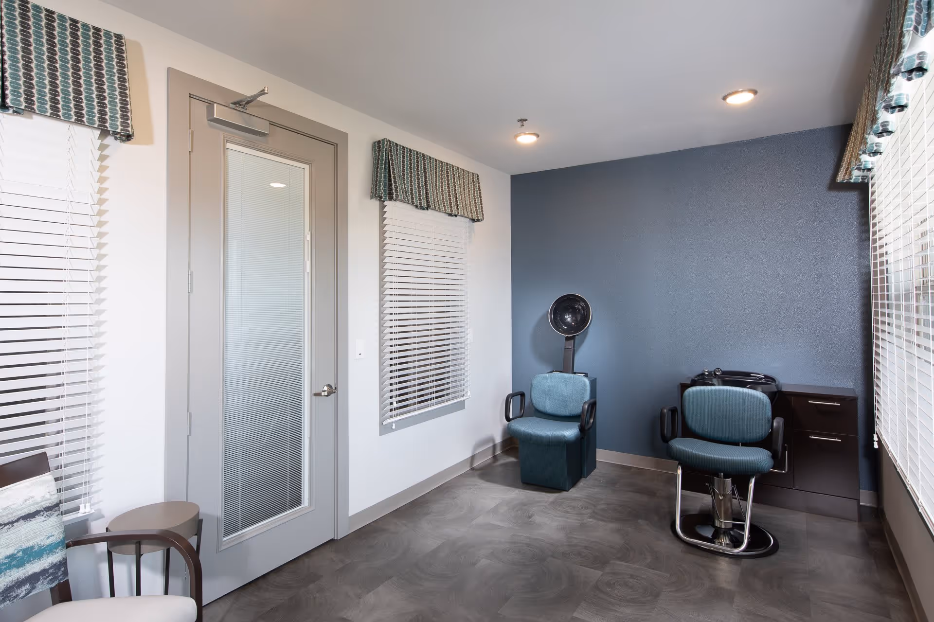 Interior view of a salon room with two teal salon chairs, one with a hair dryer hood and the other in front of a black cabinet with a sink. The room has a blue accent wall, two windows with white blinds and patterned valances, and a gray door with a glass panel.