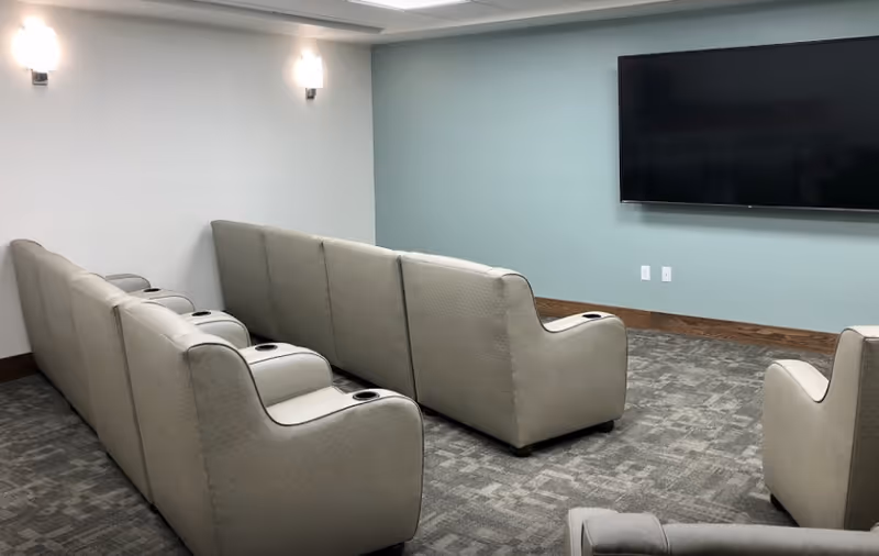 A small theater room with two rows of beige reclining seats facing a large flat-screen TV mounted on a light blue wall. The room has gray patterned carpet, white walls with two wall-mounted lights, and wooden baseboards.