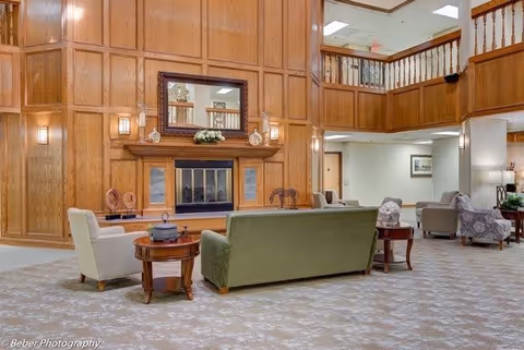 A spacious living room area with a large wooden paneled wall featuring a fireplace and a mirror above it. The room has a green sofa, white armchairs, wooden side tables, and a patterned carpet. There is a balcony railing on the upper level and soft lighting throughout the space.