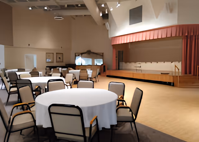 A spacious room with round tables covered in white tablecloths and surrounded by chairs. There is a small stage with a red curtain on the right side of the room. The room has high ceilings with exposed beams and soft lighting.