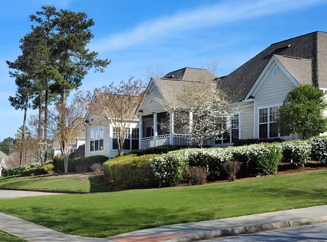 Exterior view of a single-story residential building with beige siding and a gray roof, surrounded by green grass, bushes, and trees under a clear blue sky.