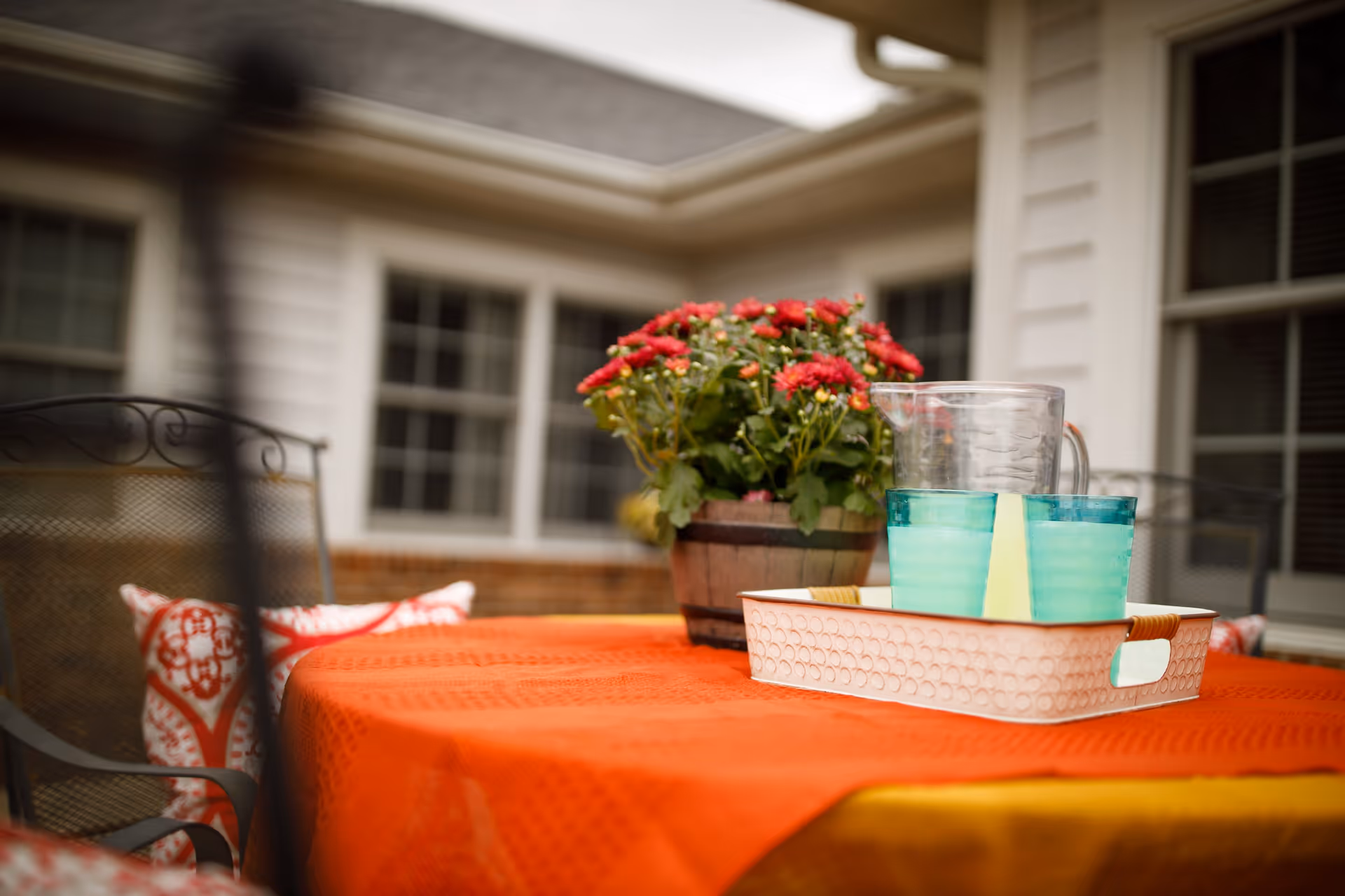 Outdoor patio table covered with an orange tablecloth, holding a white tray with a clear pitcher and two turquoise glasses, next to a pot of red flowers. The background shows windows and part of a building exterior.