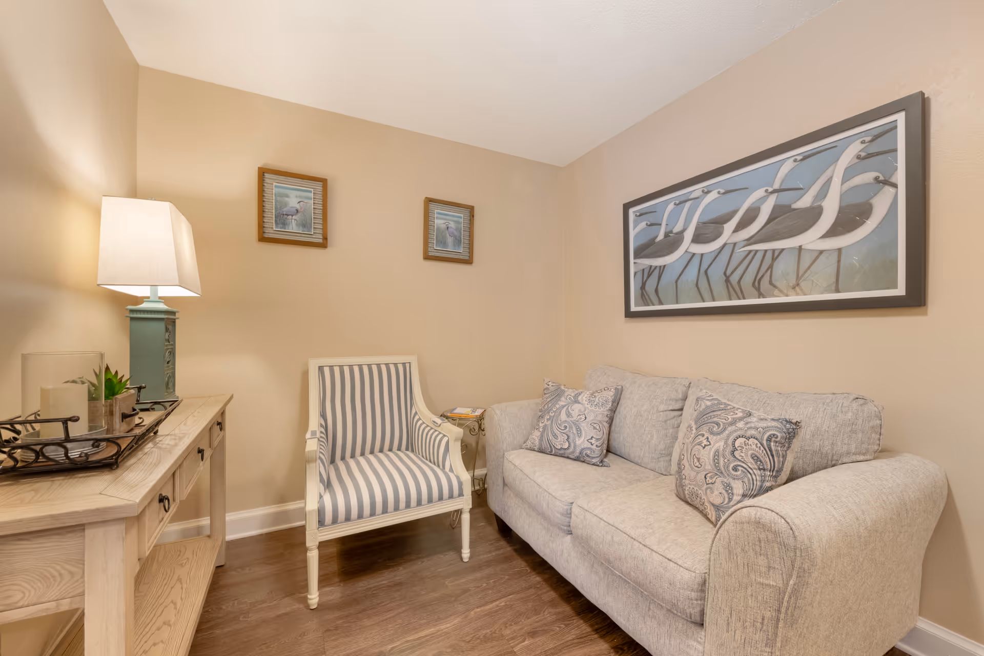 A cozy sitting area in a senior living facility featuring a light gray upholstered sofa with two patterned pillows, a striped armchair, a wooden side table with a lamp, and wall art depicting birds. The walls are painted beige and the floor is wooden.