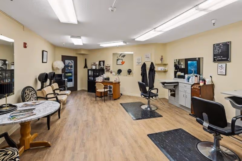 Interior view of a senior living community salon with wooden flooring, beige walls, salon chairs, hair drying stations, mirrors, and a small round table with chairs. The room is well-lit with ceiling lights and has various salon equipment and decor.