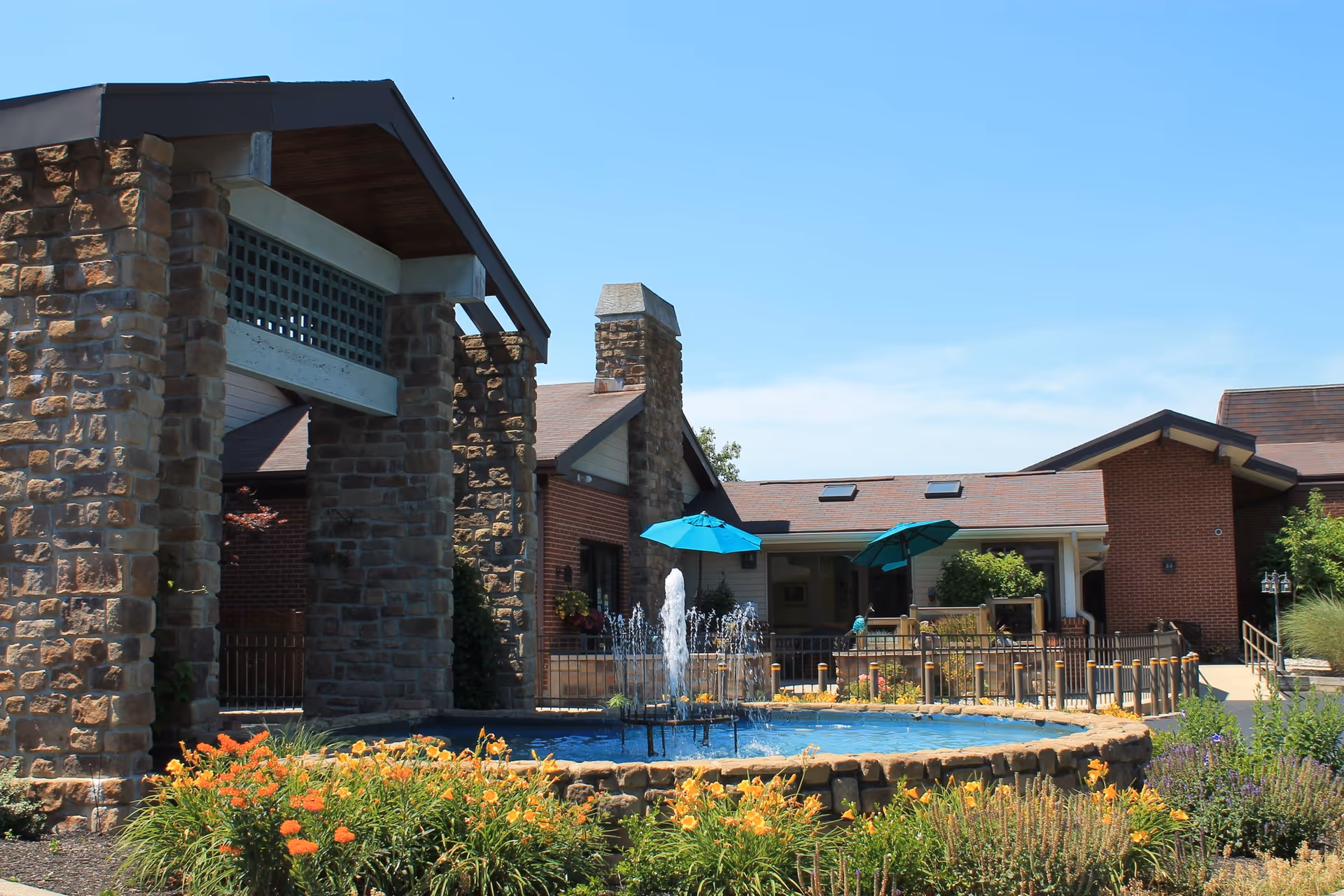 Outdoor view of a senior living facility featuring a stone and brick building with a covered entrance, a circular water fountain in the foreground, colorful flowers, and patio umbrellas providing shade over seating areas.