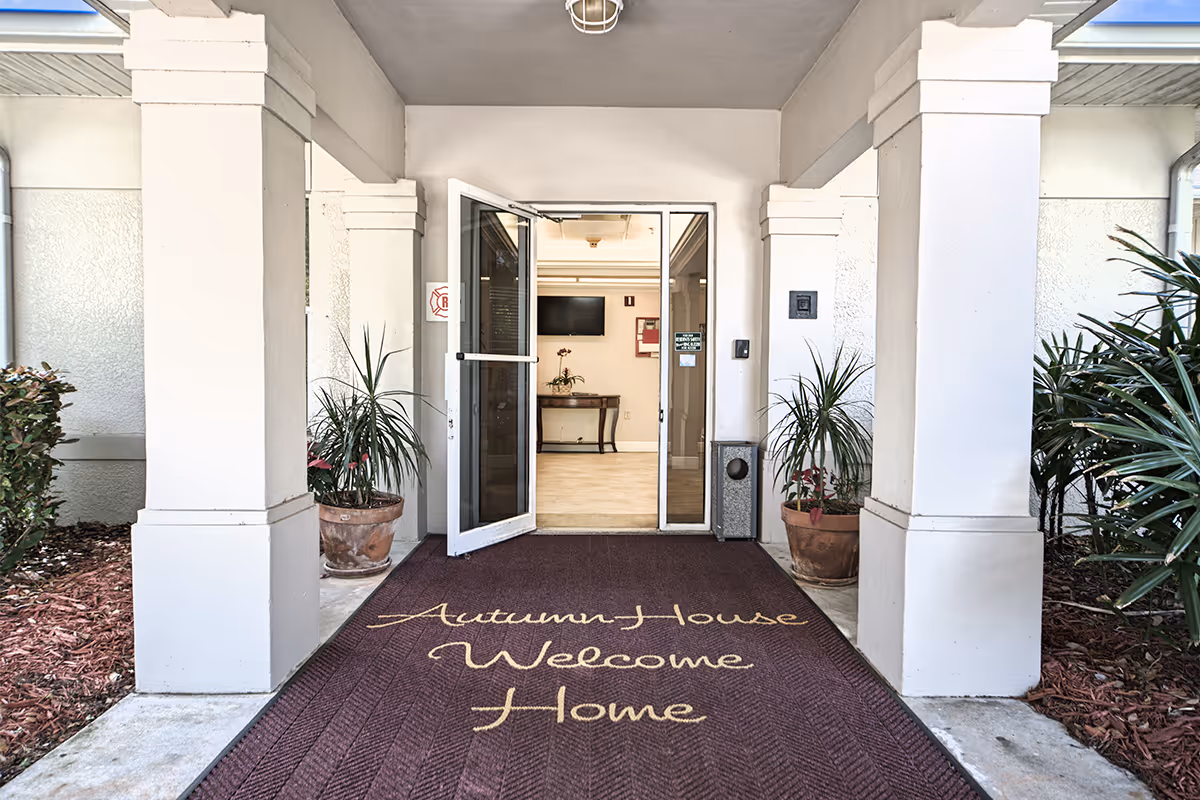 Entrance of Autumn House with columns, potted plants, and an open glass door over a welcome mat reading "Autumn House Welcome Home".