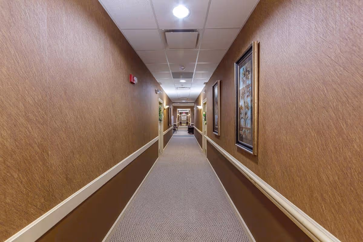 Long carpeted interior hallway with brown walls, handrails and framed artwork leading to a distant seating area.