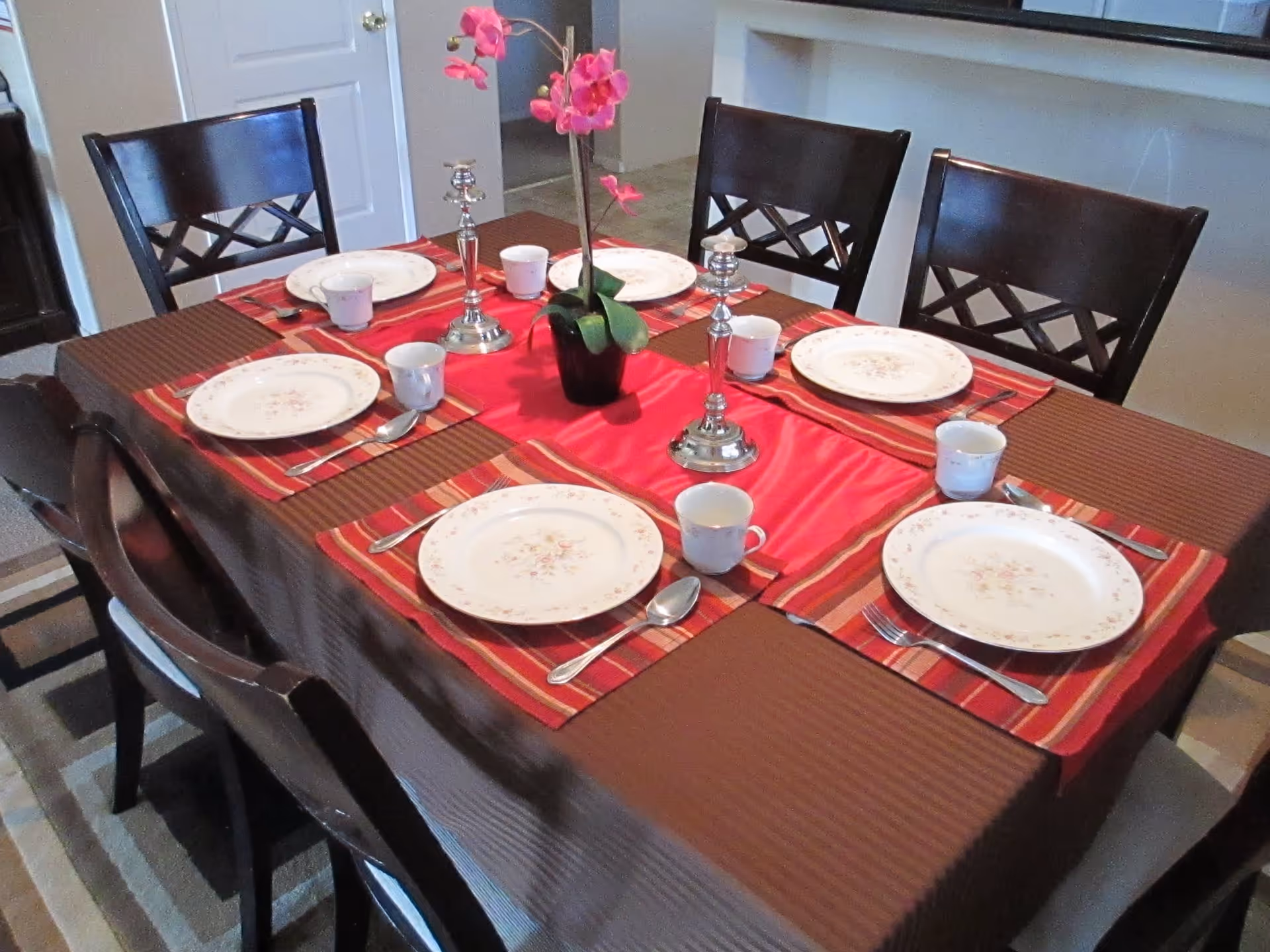 A dining table set for six with white floral plates, cups, silverware, and two silver candlesticks. The table has a brown tablecloth with red and striped placemats and a pink orchid centerpiece. Dark wooden chairs surround the table.