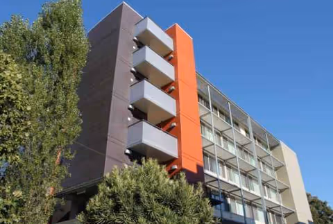 Multi-story senior living building exterior with balconies and orange accent, against blue sky.