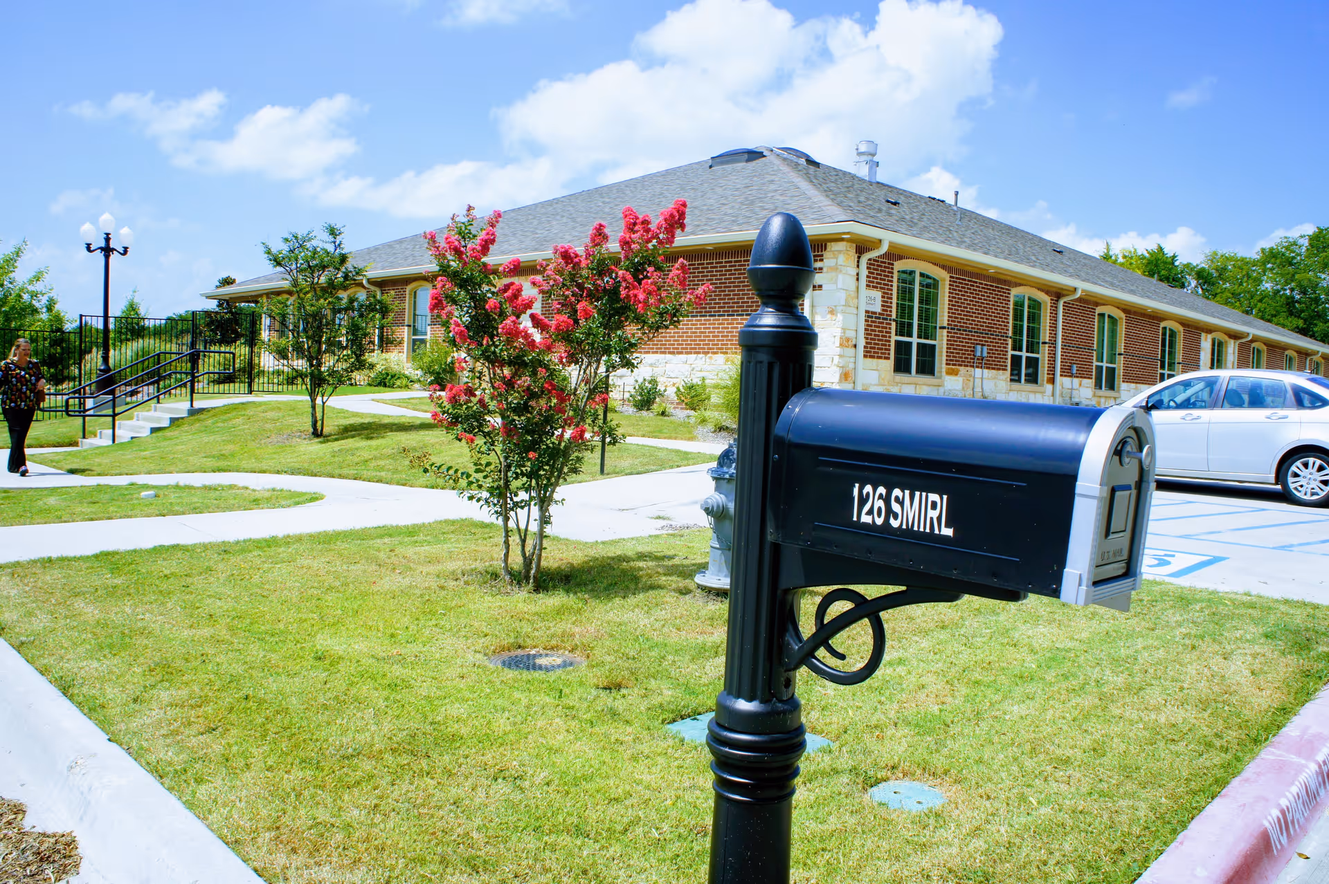 A black mailbox with the number 126 SMIRL on it stands in front of a single-story brick building with large windows. The building is surrounded by a well-maintained lawn, a flowering tree with pink blossoms, a sidewalk, and a parking area with a white car. A person is walking on the sidewalk near the building under a partly cloudy blue sky.